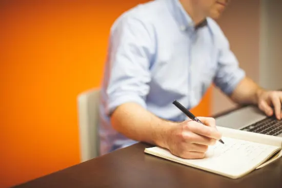 A man working at a desk takes notes with one hand while operating a computer with the other.