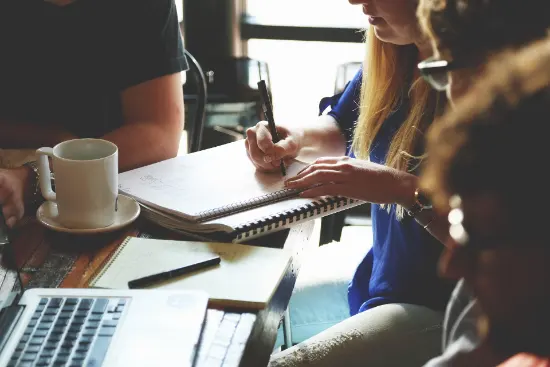 People seated at a table with coffee and a computer, a woman is taking notes.